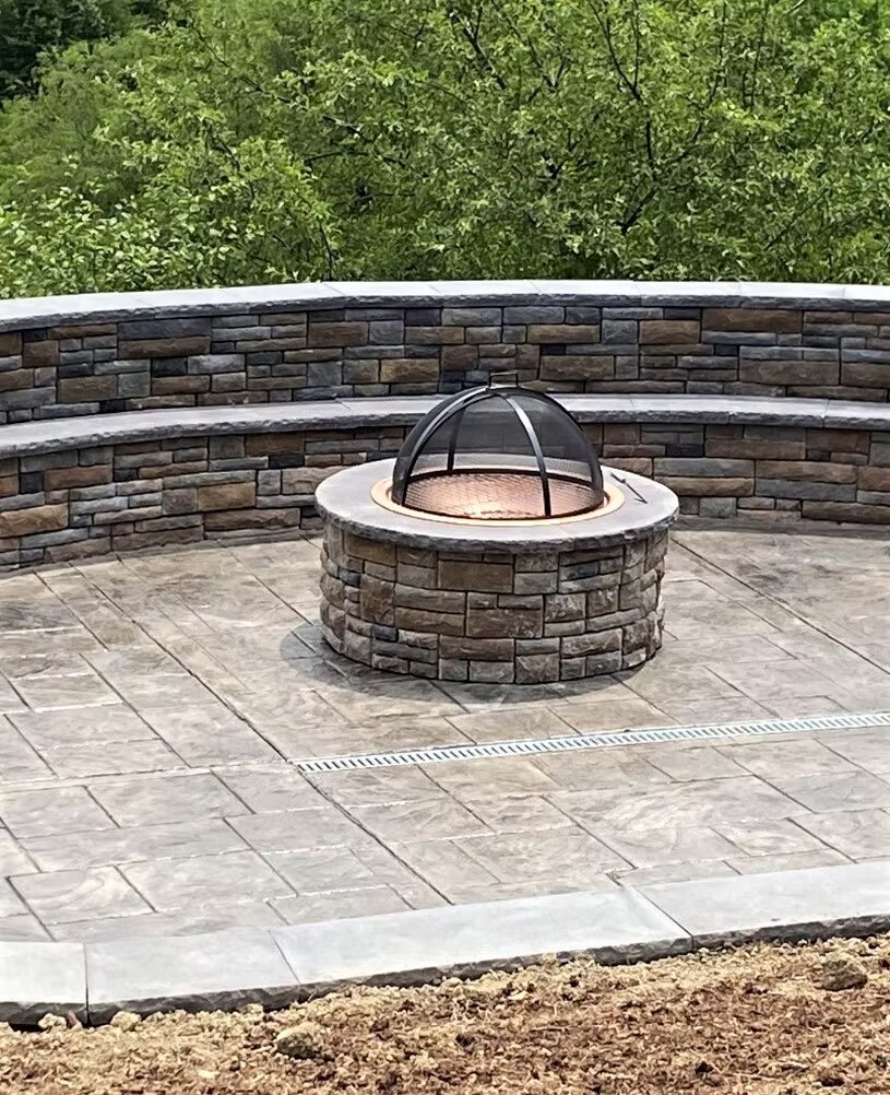 Curved stone block retaining wall and seating area surrounding a round concrete fire pit on a stamped patio